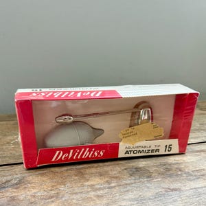 May include: A vintage DeVilbiss Atomizer 15 in its original box. The box is red and white with the brand name and product information. The atomizer is gray with a silver metal tube and adjustable tip. The box has a clear plastic window.