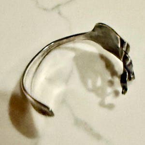 May include: A silver-toned cuff bracelet with a leaf-like design. The bracelet has a curved, open design with a textured leaf element at the top. The metal has a polished finish, reflecting light. The bracelet is designed to be worn on the wrist.