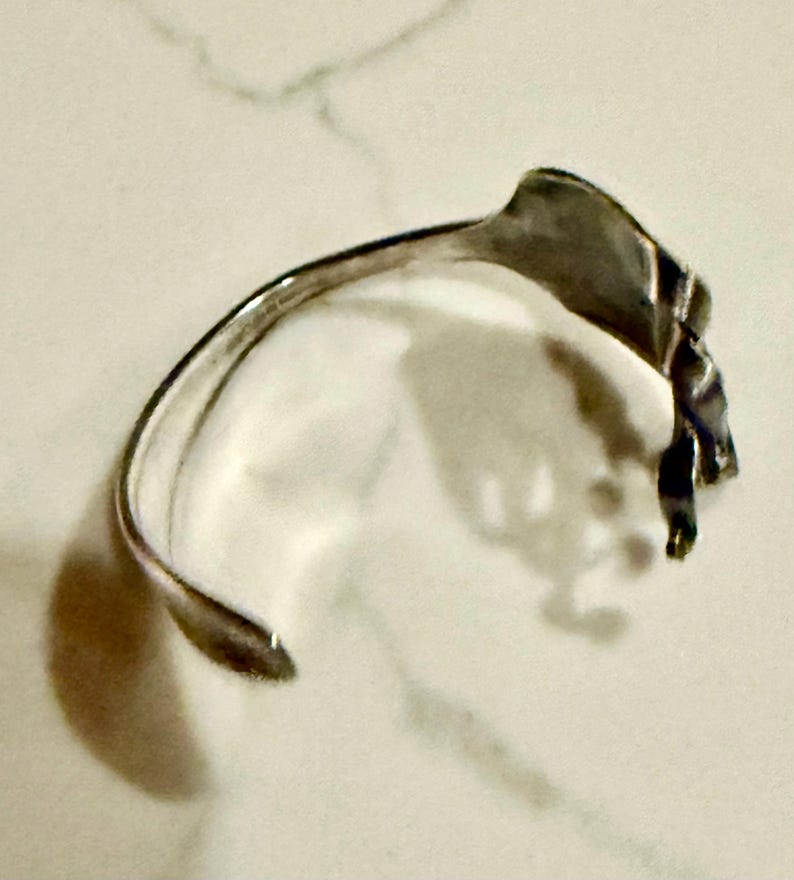 May include: A silver-toned cuff bracelet with a leaf-like design. The bracelet has a curved, open design with a textured leaf element at the top. The metal has a polished finish, reflecting light. The bracelet is designed to be worn on the wrist.