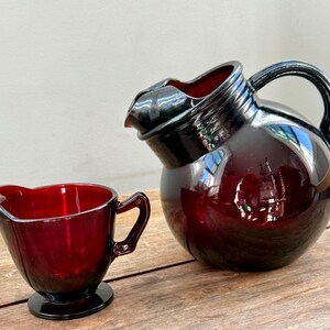 May include: A ruby red glass pitcher and creamer set. The pitcher has a rounded body, a curved handle, and a spout. The creamer has a similar design with a small handle and spout. Both pieces sit on a wooden surface.
