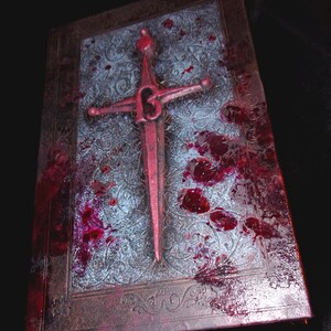 May include: A decorative book with a red sword emblem and blood splatter. The book has a silver patterned cover and a brown border. The sword is red with a stylized design. The overall theme is gothic or horror.