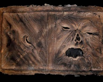 May include: A textured, brown, rectangular wall plaque with a stitched design. The plaque features a grotesque, stitched-up face with a gaping mouth and two eyes.