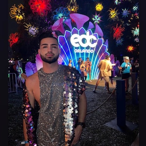 May include: A person wearing a silver sequined shirt with a purple and silver sequined shawl standing in front of a large illuminated sign that reads "edc ORLANDO".