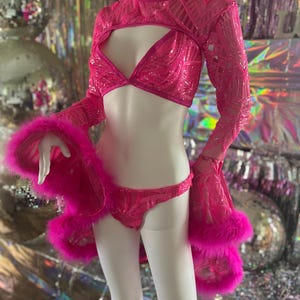 May include: A hot pink sequined lingerie set on a mannequin. The set includes a crop top with long sleeves, a bra, and a thong. The sleeves and the edges of the jacket are trimmed with pink feathers. The mannequin is set against a shiny, reflective background.