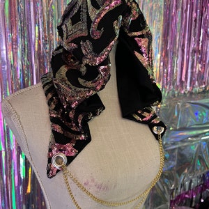 May include: A black sequined scarf with pink and gold accents, featuring a chain with two metal rings. The scarf is draped over a white mannequin.