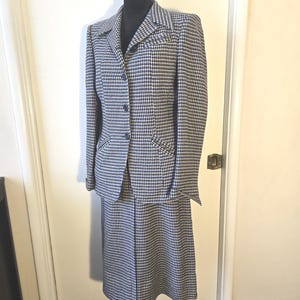 May include: A vintage houndstooth skirt suit in black, white, and navy blue. The jacket has a notched collar, three buttons, and two front pockets. The skirt is knee-length. The suit is displayed on a black mannequin.