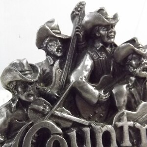 K&ouml;nnte beinhalten: Silberne G&uuml;rtelschnalle mit vier Musikern, die Cowboyh&uuml;te tragen und Instrumente spielen. Die Schnalle tr&auml;gt die Aufschrift "Country" in gro&szlig;en Buchstaben.