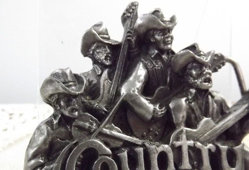 K&ouml;nnte beinhalten: Silberne G&uuml;rtelschnalle mit vier Musikern, die Cowboyh&uuml;te tragen und Instrumente spielen. Die Schnalle tr&auml;gt die Aufschrift "Country" in gro&szlig;en Buchstaben.