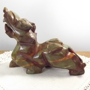 May include: A green and brown jade carving of a dragon with its head tilted back. The dragon is lying on its side with its tail curled up.