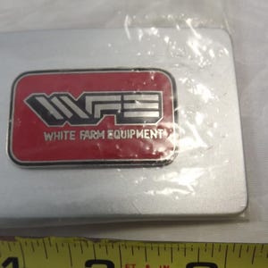 May include: A silver metal belt buckle with a red rectangular logo that says "WFE White Farm Equipment" in white letters.