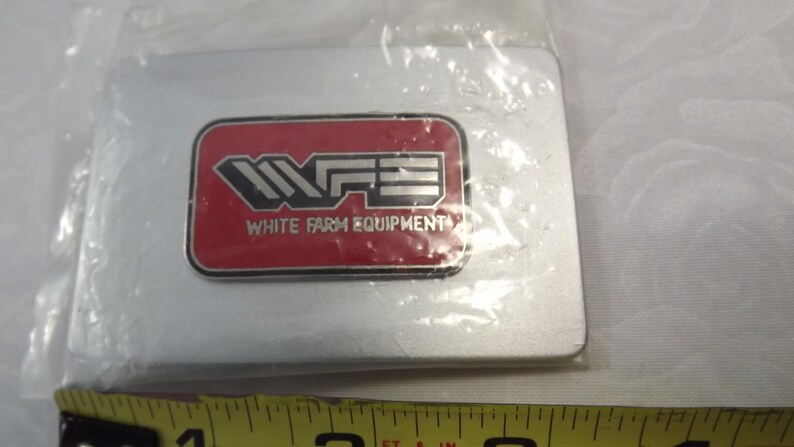 May include: A silver metal belt buckle with a red rectangular logo that says "WFE White Farm Equipment" in white letters.