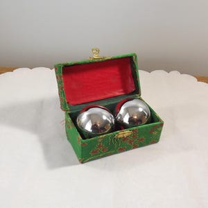 May include: Two polished silver-coloured metal spheres are nestled inside a rectangular green fabric-covered box. The box features a floral pattern, a gold-toned metal latch, and a vibrant red interior lining that contrasts with the metallic spheres.