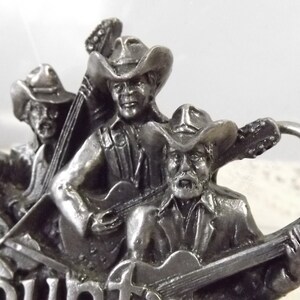 K&ouml;nnte beinhalten: Silberne G&uuml;rtelschnalle mit vier Cowboys, die Musikinstrumente spielen. Die Schnalle tr&auml;gt die Gravur "Country" am unteren Rand.