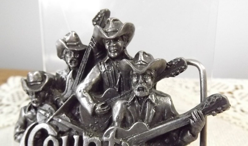 K&ouml;nnte beinhalten: Silberne G&uuml;rtelschnalle mit vier Cowboys, die Musikinstrumente spielen. Die Schnalle tr&auml;gt die Gravur "Country" am unteren Rand.