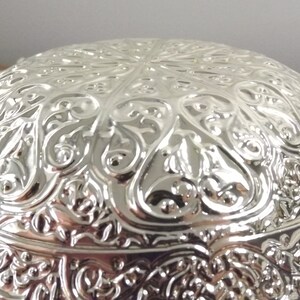 Puede incluir: Un cuenco de plata con un dise&ntilde;o ornamentado y en relieve. El cuenco tiene forma redonda y una superficie lisa y reflectante.