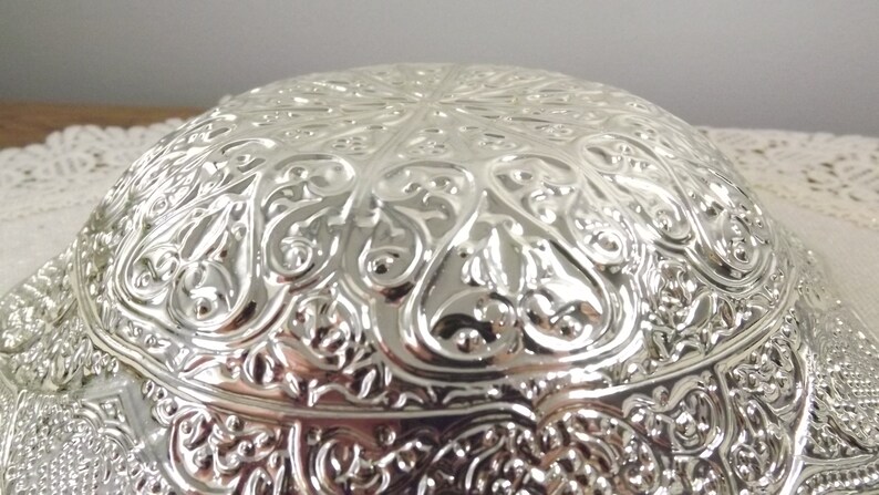 Puede incluir: Un cuenco de plata con un dise&ntilde;o ornamentado y en relieve. El cuenco tiene forma redonda y una superficie lisa y reflectante.