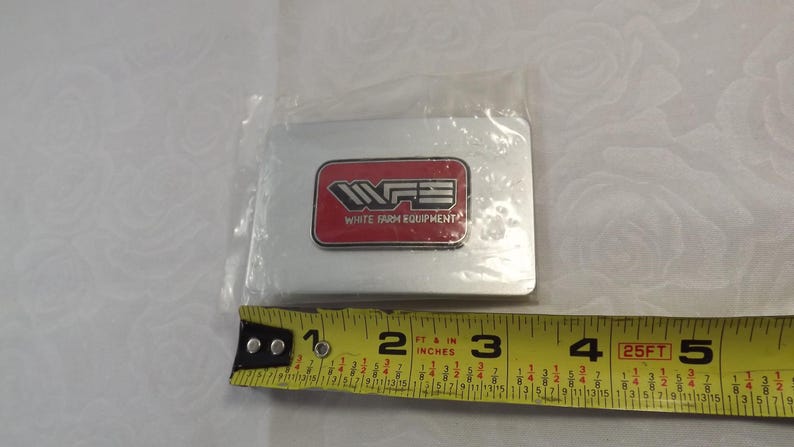 May include: A silver metal rectangle with a red rectangle inside. The red rectangle has the text "WFE" in white and the text "WHITE FARM EQUIPMENT" in black.
