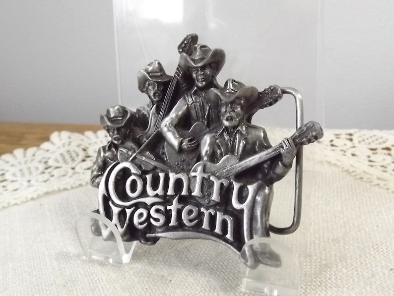K&ouml;nnte beinhalten: Silberne G&uuml;rtelschnalle mit einer Gruppe von vier Musikern, die Instrumente spielen. Die Schnalle tr&auml;gt die Aufschrift "Country Western".
