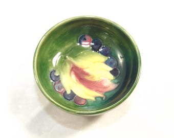 May include: A small green ceramic bowl with a painted design of a yellow and pink leaf with purple and blue berries.