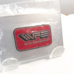 May include: A red and silver metal pin with the letters "WFE" and the text "White Farm Equipment".