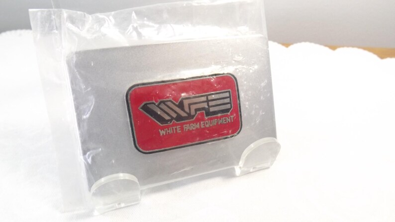 May include: A red and silver metal pin with the letters "WFE" and the text "White Farm Equipment".