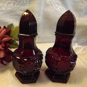 May include: Two antique ruby red glass salt and pepper shakers. The shakers have a decorative design and a faceted top. The base is square and the shakers are displayed on a white surface.