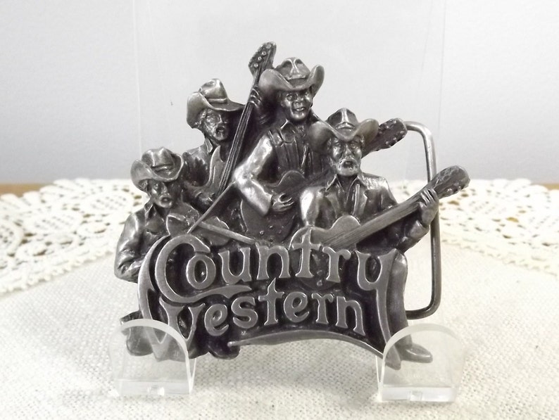 K&ouml;nnte beinhalten: Silberne G&uuml;rtelschnalle mit einer Gruppe von vier Country-Musikern, die Instrumente spielen. Die Schnalle ist mit den Worten "Country Western" graviert.