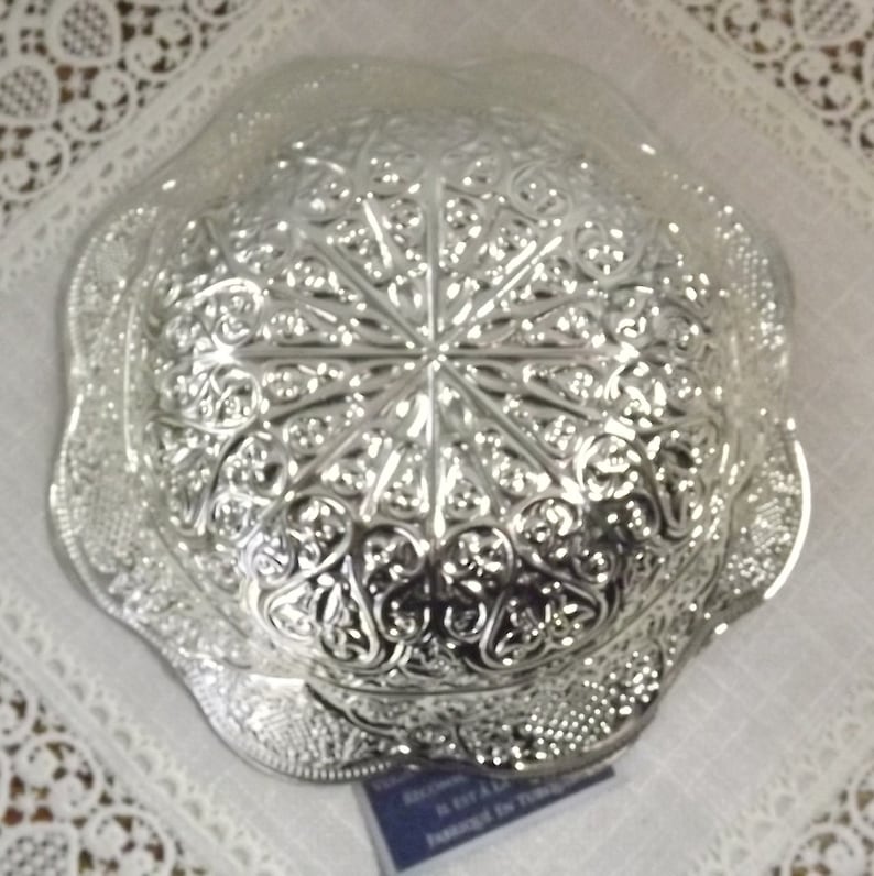 Puede incluir: Un cuenco de plata con un dise&ntilde;o intrincado en relieve. El cuenco tiene un borde festoneado y una base circular.