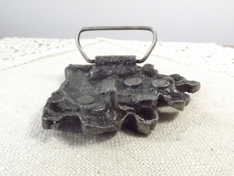 K&ouml;nnte beinhalten: Eine silberfarbene G&uuml;rtelschnalle mit einem detaillierten Design einer Bergkette. Die Schnalle hat eine einfache, rechteckige Schlaufe zum Befestigen an einem G&uuml;rtel.