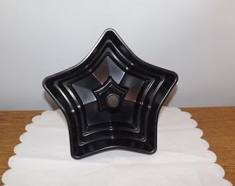 May include: A black star-shaped baking pan with a smooth, metallic finish. The pan features a distinct five-pointed star design with tiered, concentric ridges leading toward a central circular opening, creating a geometric, multi-dimensional look.
