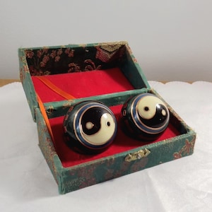 May include: Two black and white Yin Yang exercise balls presented in a decorative box. The balls feature a white Yin Yang symbol on a black surface, with blue and gold detailing. The box is green with a red interior and a gold clasp.