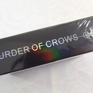 May include: A black box with the words "Murder of Crows" printed in rainbow iridescent letters. The box is wrapped in clear plastic.