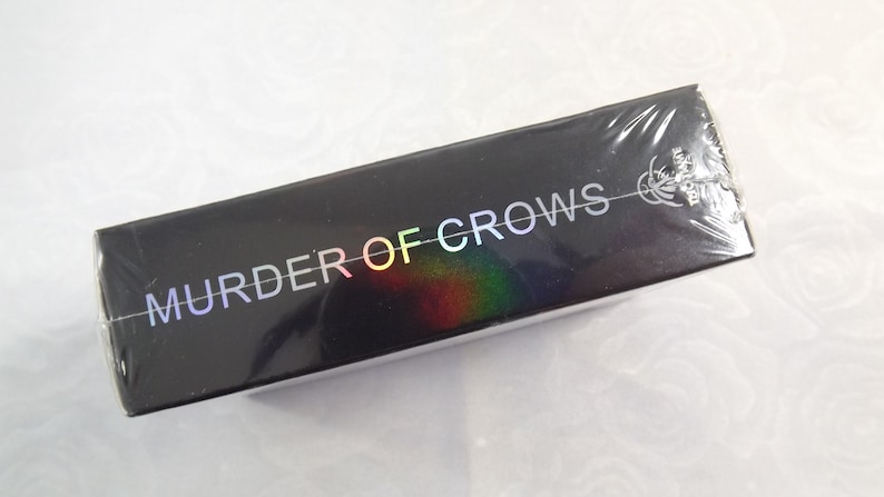 May include: A black box with the words "Murder of Crows" printed in rainbow iridescent letters. The box is wrapped in clear plastic.