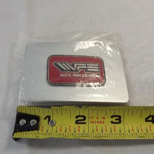 May include: A silver metal rectangular sign with a red rectangle in the center. The red rectangle has the text "WFE" in white letters and the text "White Farm Equipment" in smaller white letters.
