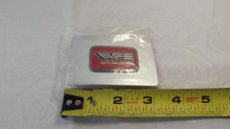 May include: A silver metal rectangular sign with a red rectangle in the center. The red rectangle has the text "WFE" in white letters and the text "White Farm Equipment" in smaller white letters.