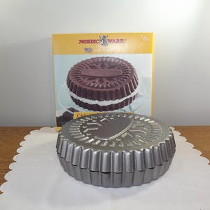 Peut inclure: Un moule à pâtisserie en métal argenté, cannelé, en forme de grand biscuit sandwich, avec une boîte assortie derrière. La boîte présente une image du biscuit fini, avec les mots "Nordic Ware" en haut.
