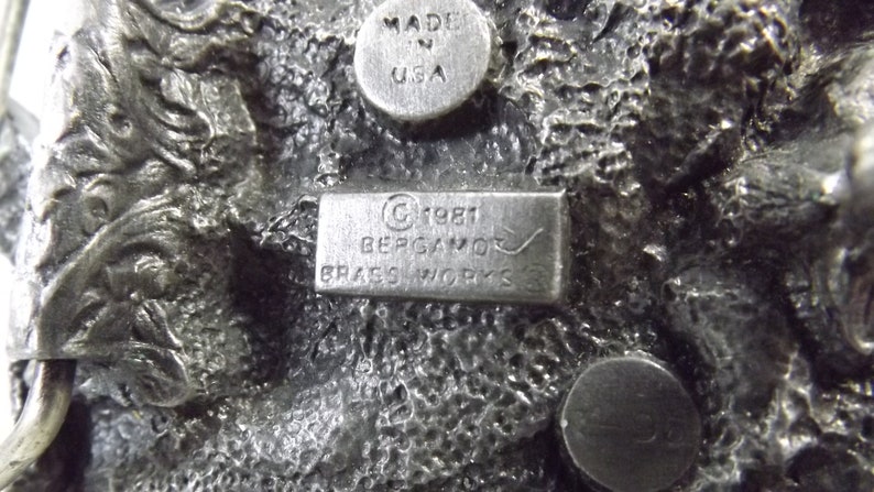 K&ouml;nnte beinhalten: Eine silberne G&uuml;rtelschnalle mit einem detaillierten Design und dem Text "C 1981 BERGAMO BRASS WORKS" auf der R&uuml;ckseite.