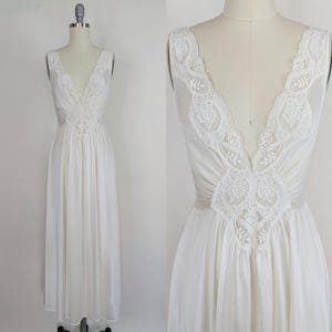 Puede incluir: Un camisón vintage blanco con un escote en V profundo y delicados adornos de encaje. El camisón sin mangas presenta intrincados detalles de encaje en el corpiño y una falda larga y fluida. El vestido se exhibe en un maniquí.