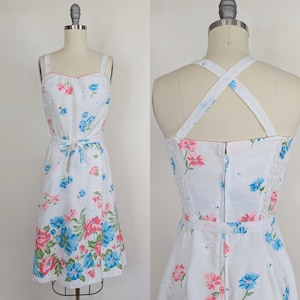 May include: White sundress with a floral pattern of pink and blue flowers. The dress has a square neckline with pink trim, a tie waist, and a zipper closure in the back. The straps cross in the back. The dress is displayed on a mannequin.