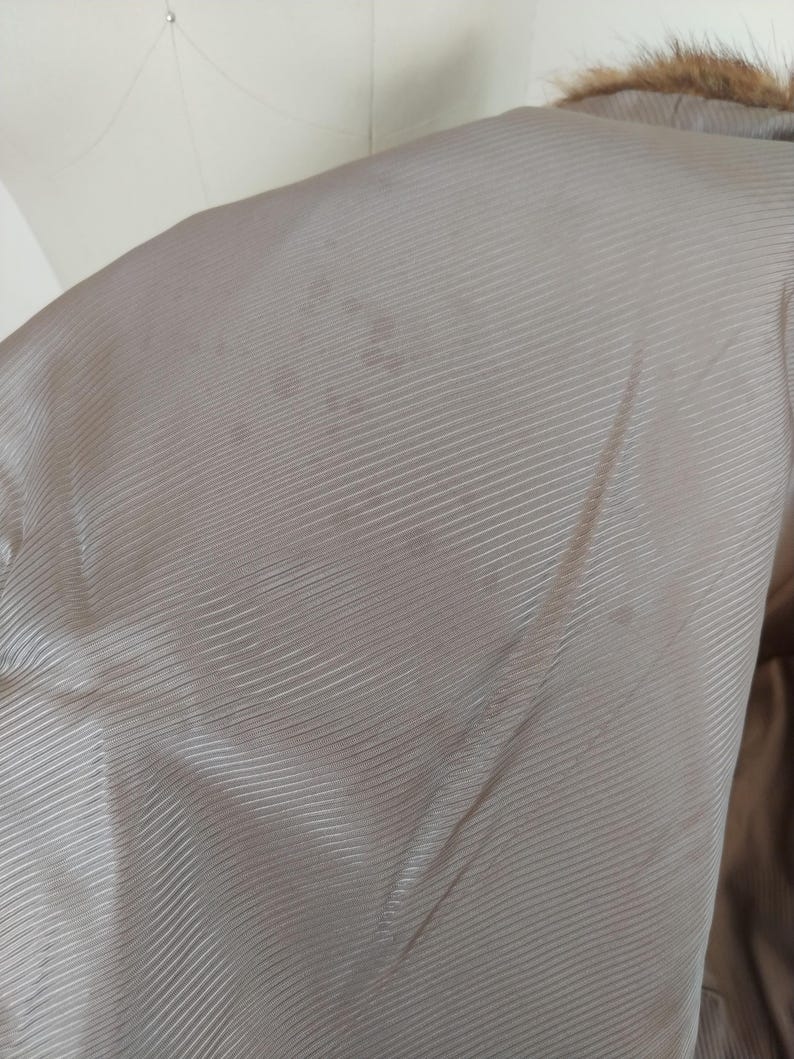 May include: Close-up of the interior lining of a coat. The lining is a light tan color with a subtle, ribbed texture. The coat's fur collar is visible at the top. There are some darker spots on the lining.