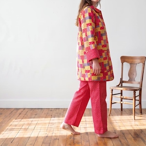 May include: A long-sleeved jacket featuring a multi-colored patchwork pattern in shades of red, yellow, and blue, paired with matching red trousers that have a small white geometric print. The jacket has a collar and long sleeves with cuffs.