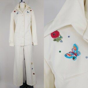 Peut inclure: Un tailleur blanc compos&eacute; d'une veste &agrave; manches longues boutonn&eacute;e et d'un pantalon &agrave; jambes larges. Le tailleur est d&eacute;cor&eacute; de fleurs et de papillons brod&eacute;s dans des tons de rouge, bleu, vert et jaune.