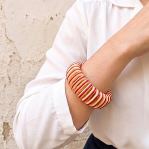 May include: A chunky bangle bracelet featuring a repeating pattern of alternating thick cream-colored and thin red circular rings. The bracelet has a coiled, spring-like structure with a smooth finish, creating a bold, layered appearance on the wrist.