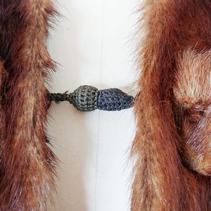 May include: Close-up of a vintage fur coat with a dark brown and reddish-brown color. The coat features a decorative clasp in shades of green and gray. The fur appears soft and luxurious, with a detailed texture.