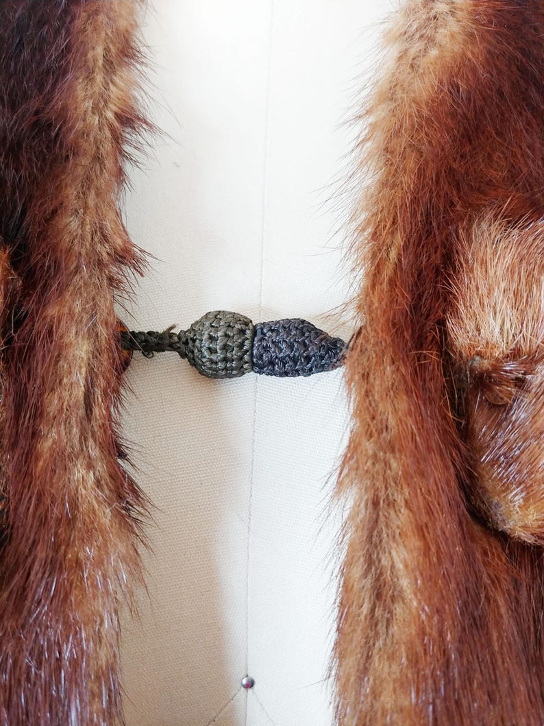 May include: Close-up of a vintage fur coat with a dark brown and reddish-brown color. The coat features a decorative clasp in shades of green and gray. The fur appears soft and luxurious, with a detailed texture.