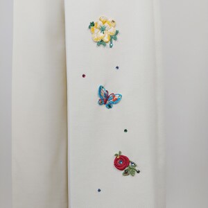 Peut inclure: Tissu blanc avec des fleurs brod&eacute;es et un papillon. Les fleurs sont jaunes, rouges et vertes. Le papillon est bleu et vert. Il y a aussi de petits strass clairs &eacute;parpill&eacute;s sur le tissu.
