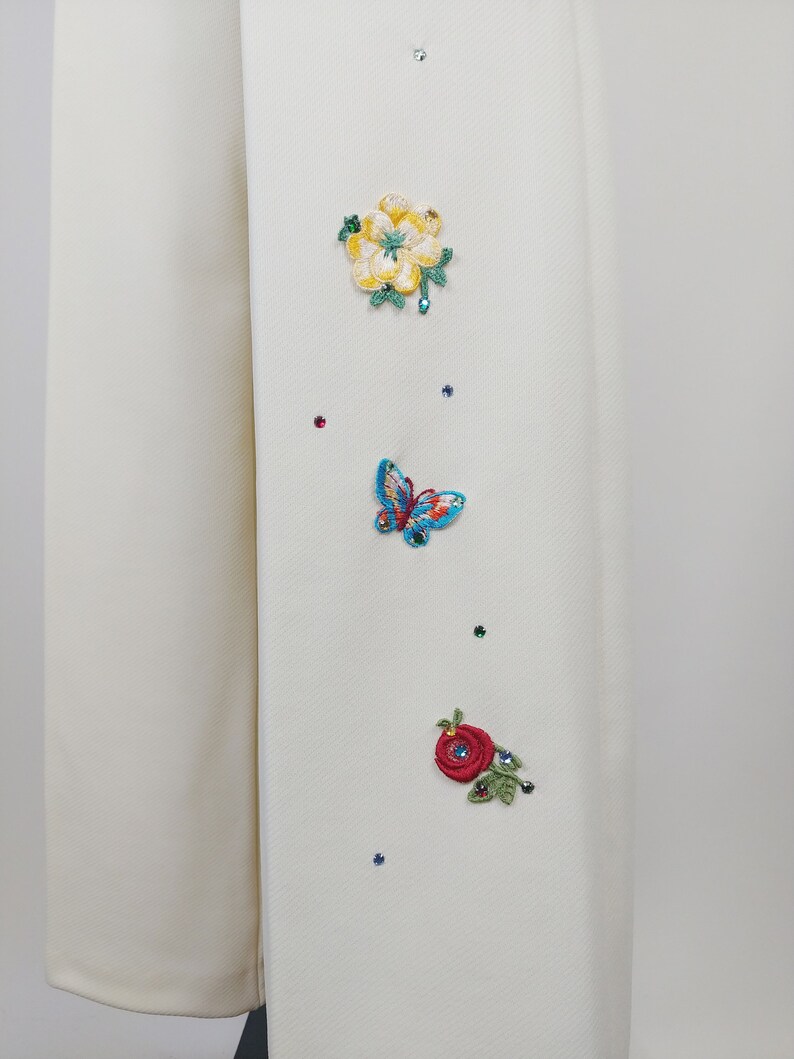 Peut inclure: Tissu blanc avec des fleurs brod&eacute;es et un papillon. Les fleurs sont jaunes, rouges et vertes. Le papillon est bleu et vert. Il y a aussi de petits strass clairs &eacute;parpill&eacute;s sur le tissu.