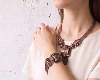 May include: A copper-toned metal necklace and matching bracelet set featuring a fine mesh chain design. Both pieces include decorative rectangular centre elements with intricate scrollwork patterns and dangling fringe accents at the bottom.