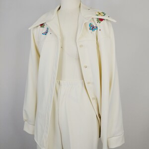 Peut inclure: Un tailleur-pantalon blanc compos&eacute; d'une veste &agrave; manches longues et d'un pantalon assorti. La veste a un col large avec des papillons et des fleurs brod&eacute;s dans des tons de rouge, bleu et vert.