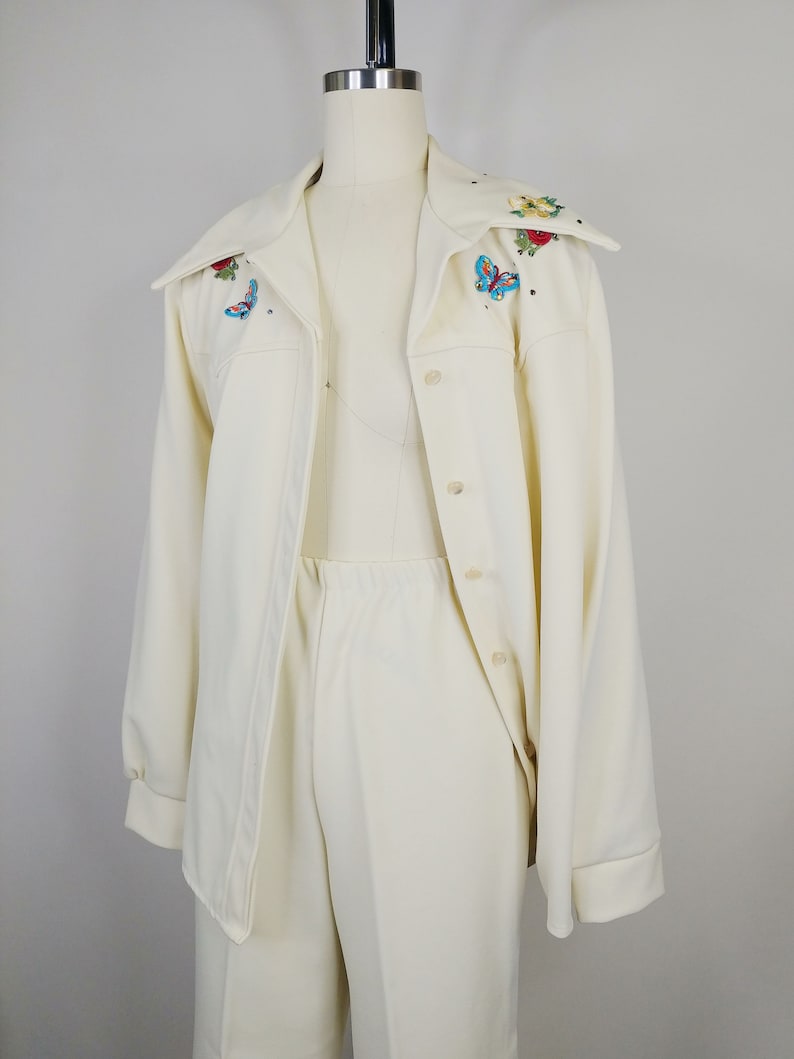 Peut inclure: Un tailleur-pantalon blanc compos&eacute; d'une veste &agrave; manches longues et d'un pantalon assorti. La veste a un col large avec des papillons et des fleurs brod&eacute;s dans des tons de rouge, bleu et vert.